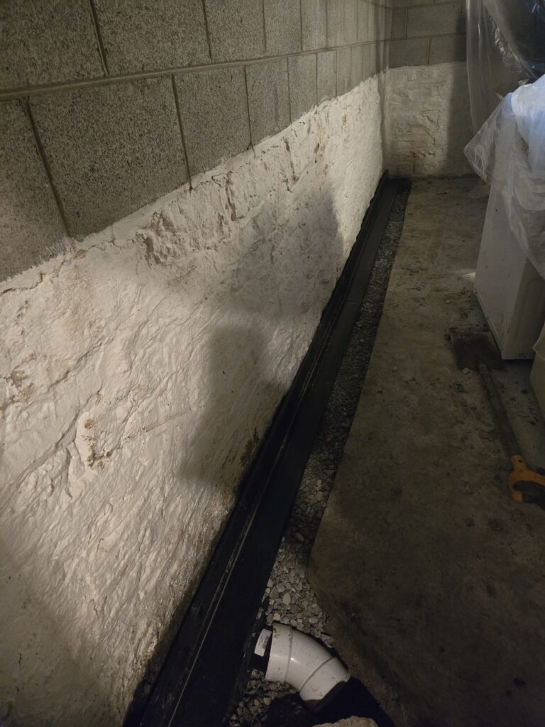 Basement with exposed concrete walls, a drainage system along the floor, and a white pipe fitting.