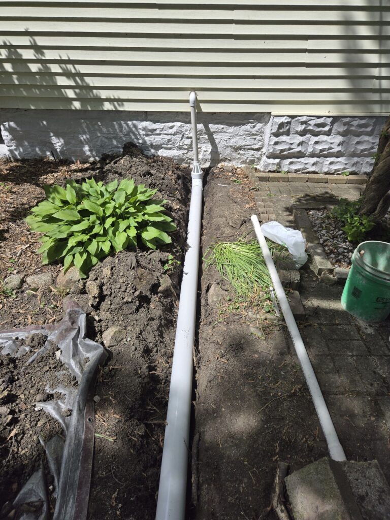 A white pipe is laid in a narrow trench beside a house. A pile of soil and a green plant are visible next to the trench.