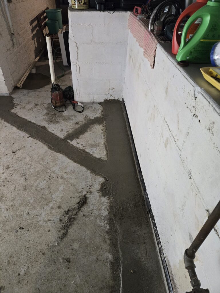 Freshly applied concrete patchwork on a garage floor corner near a brick wall, with tools and supplies visible on a shelf.