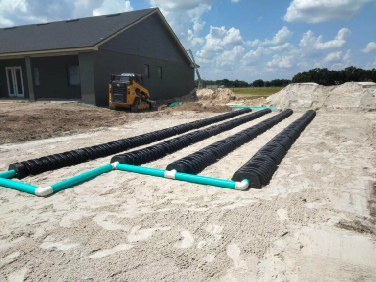 Rows of black drainage pipes with turquoise connectors laid out on sandy ground near a house under construction.