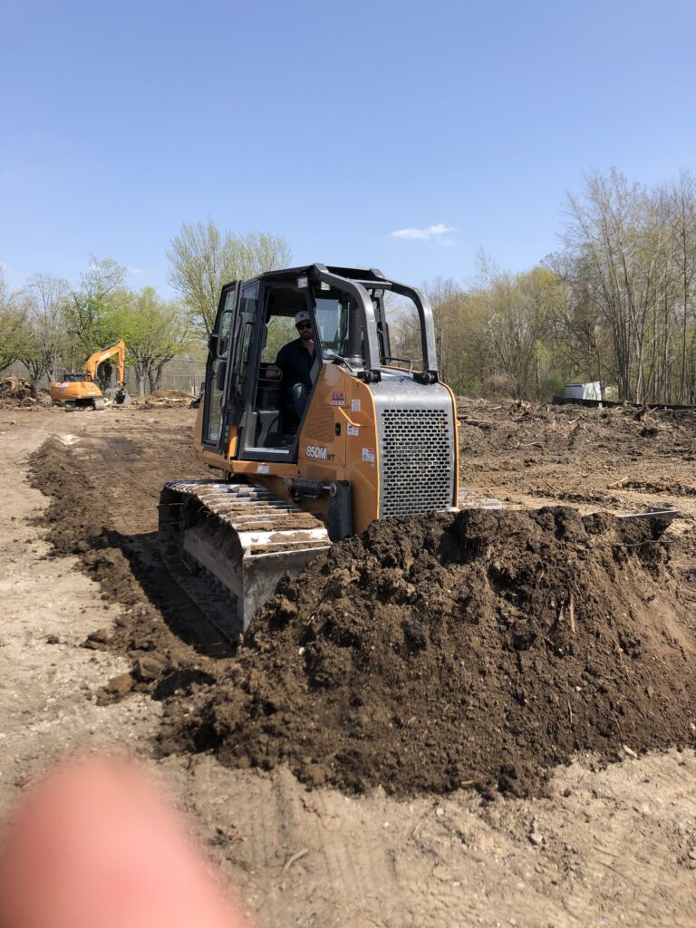 A bulldozer clearing soil on a construction site with trees in the background. Another construction vehicle is visible in the distance.