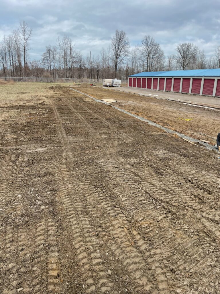 Cleared plot of land with visible tire tracks, adjacent to a row of red storage units under a cloudy sky. Bare trees line the background.