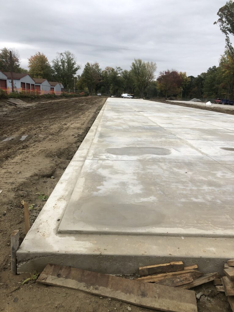 A newly constructed concrete walkway in a residential area with mud on the sides and trees in the background.