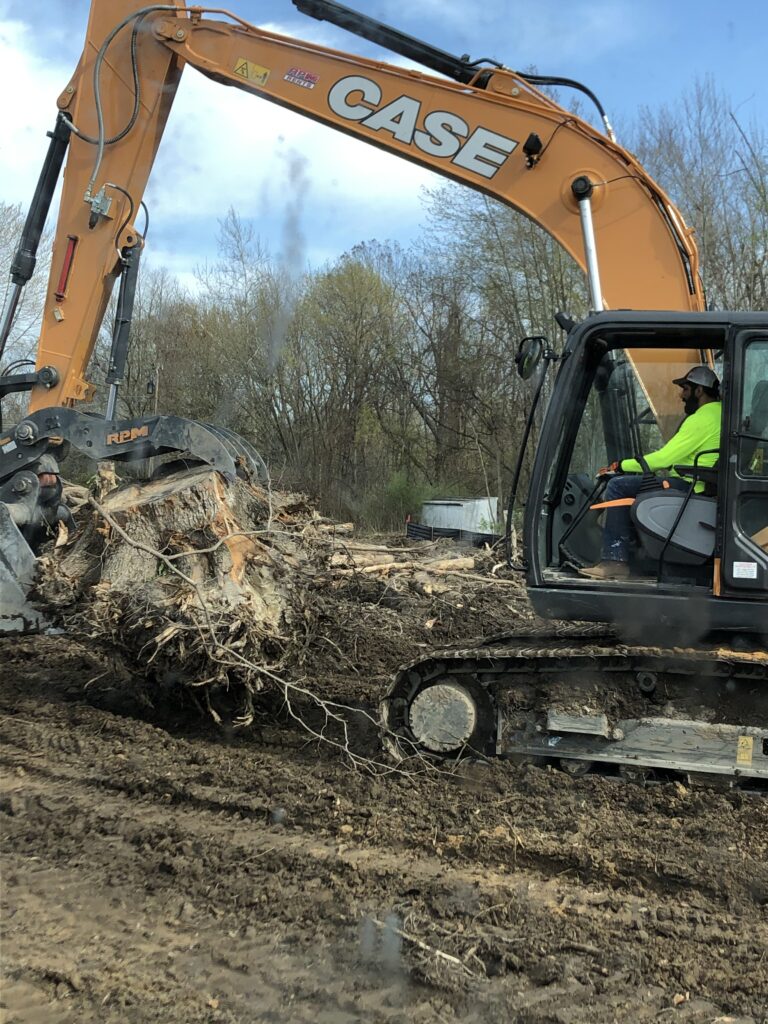 A construction worker operates an excavator to remove tree roots from a muddy construction site.