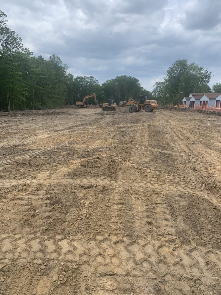 Construction site with several excavators on a cleared, dirt area surrounded by trees and a few small buildings in the background. Overcast sky above.